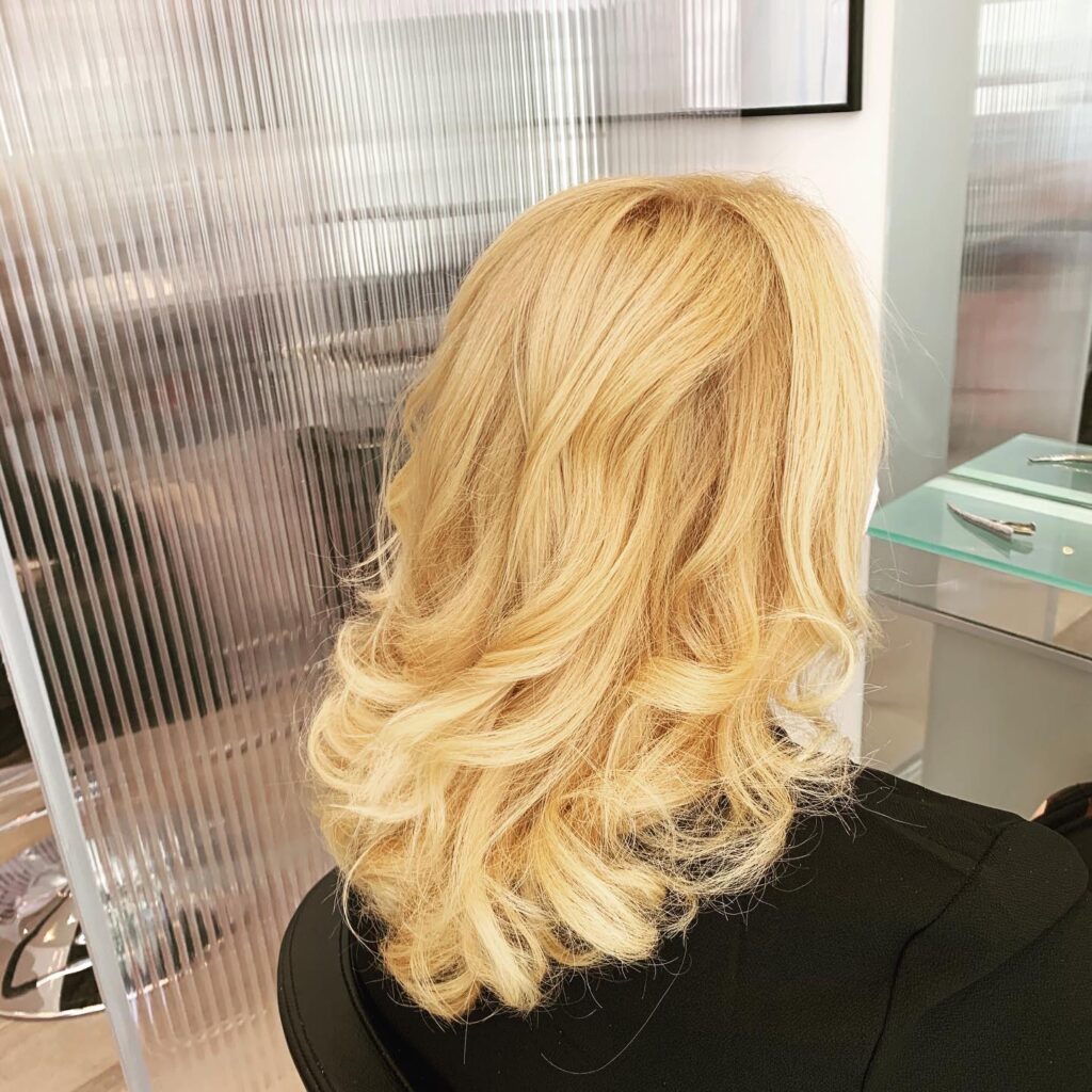 Femme de dos avec des cheveux blonds ondulés mi-longs dans un salon de coiffure moderne et lumineux.