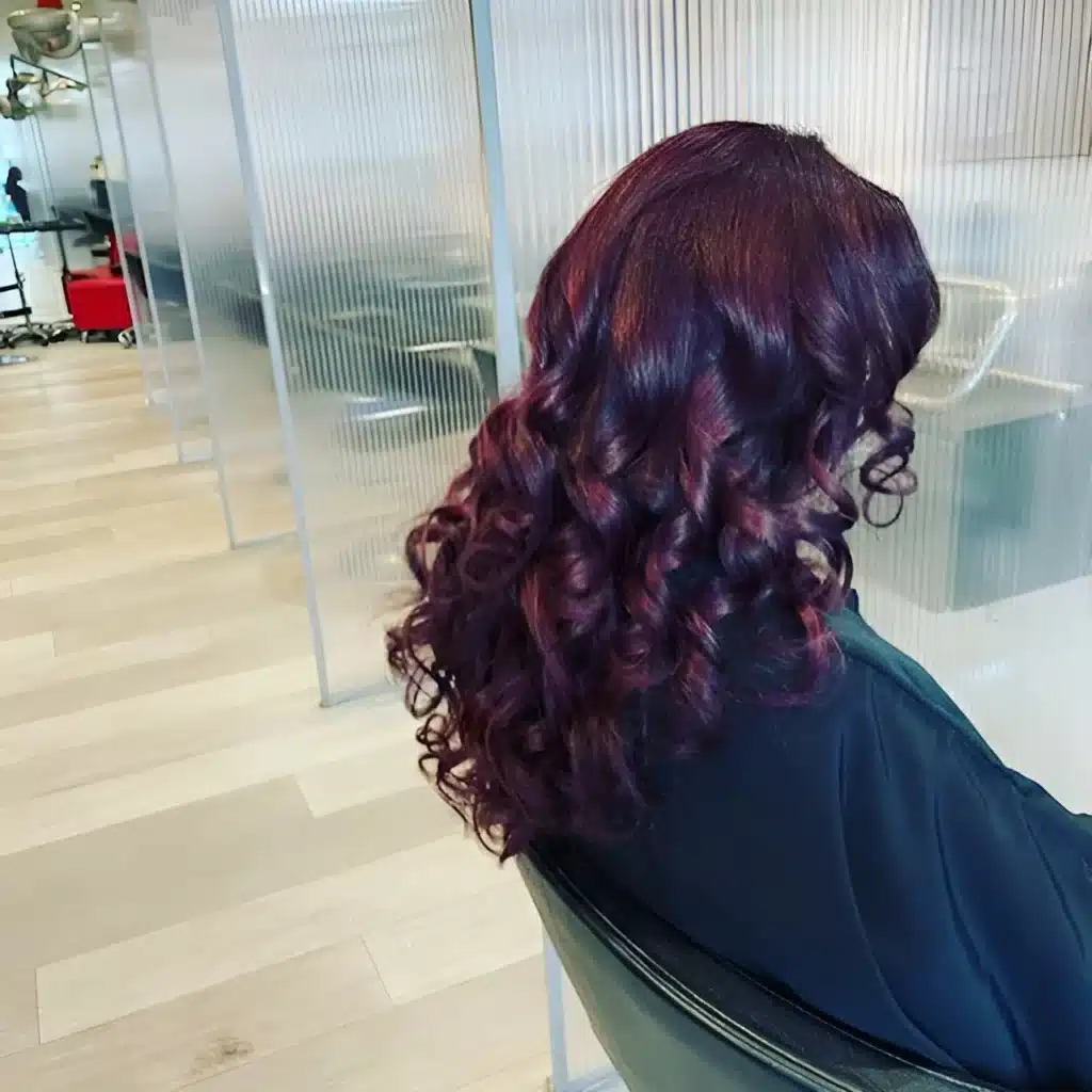 Cheveux longs bouclés couleur prune intense et reflets violets, coiffure volumineuse en salon de coiffure.