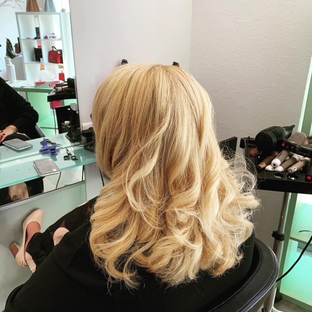 Femme aux longs cheveux blonds ondulés dans un salon de coiffure, reflet d'outils de stylisme dans le miroir.
